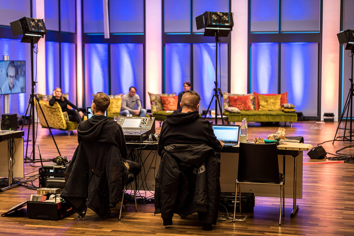 Zwei Mitarbeiter sitzen am Tisch mit Technik, im Hintergrund sitzen drei Referenten auf den Sofas