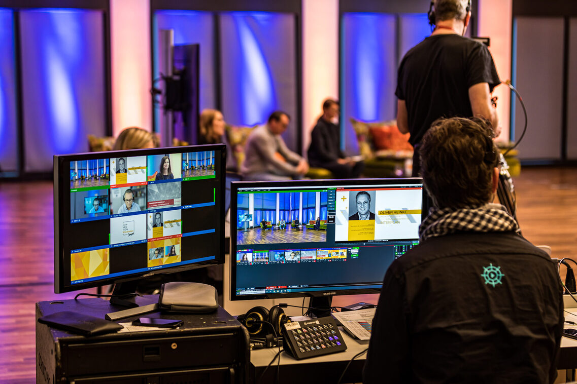 Ein Mitarbeiter steuert den Live-Stream vor dem Rechner, im Hintergrund sitzen drei Referenten