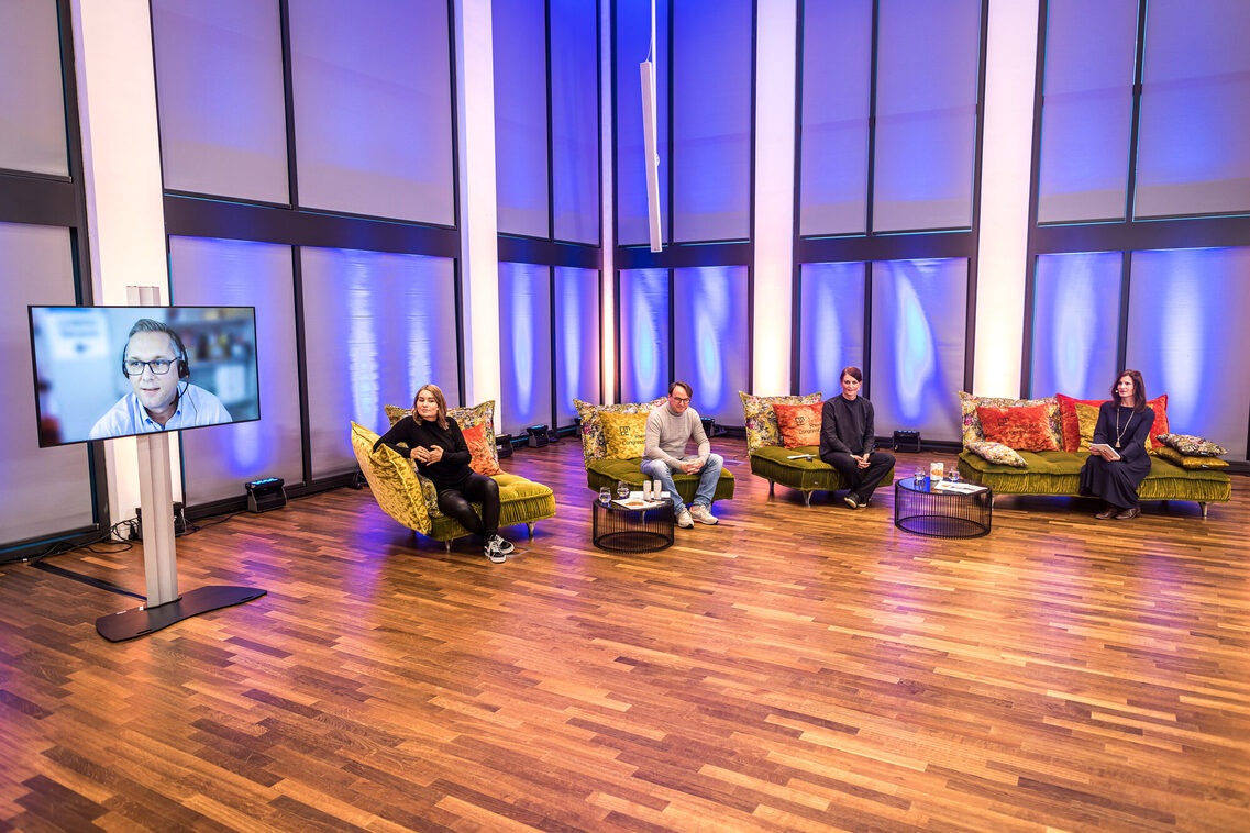 Drei Referenten und eine Moderatorin sitzen am Sofa im Studio und hören den Online-Vortrag eines vierten Referenten zu