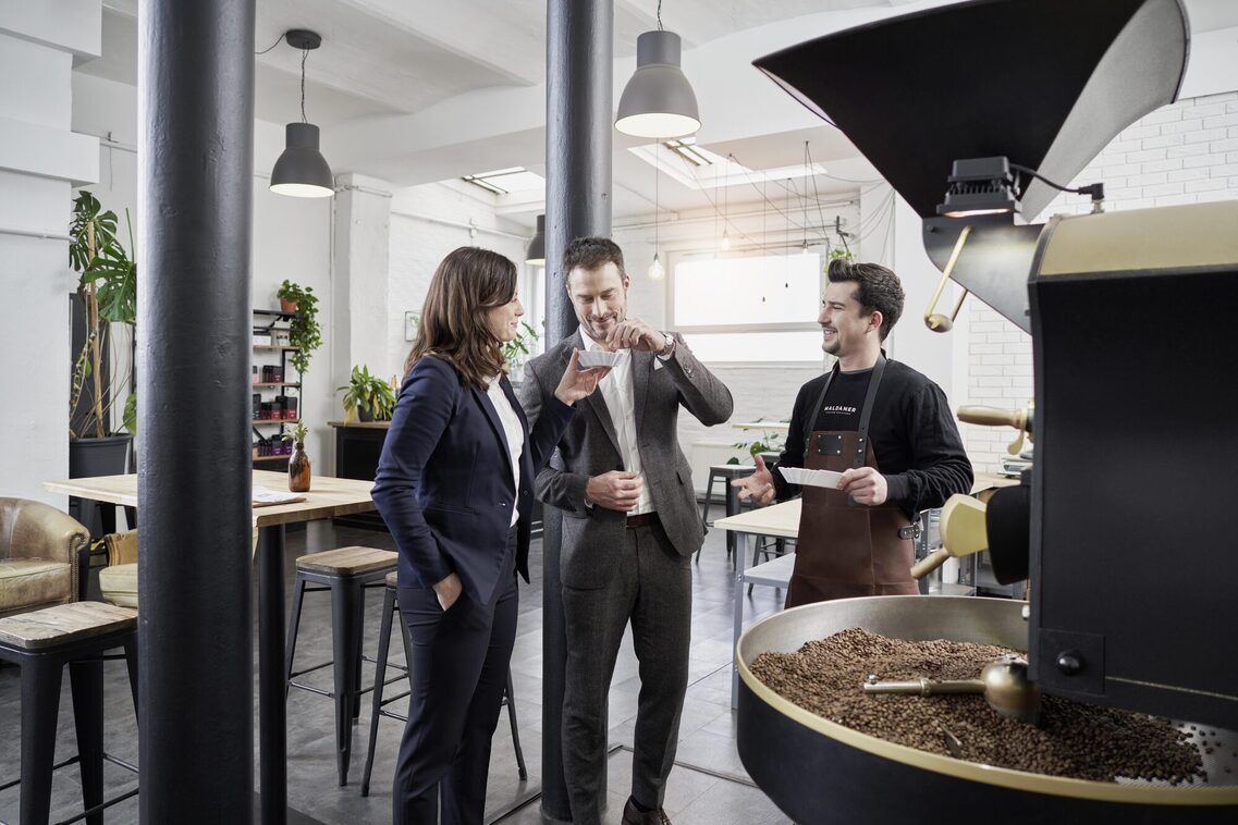 Eine Frau und ein Mann in Businesskleidung stehen vor der Kaffeeröstmaschine und ein Mitarbeiter erzählt über Kaffeebohnen