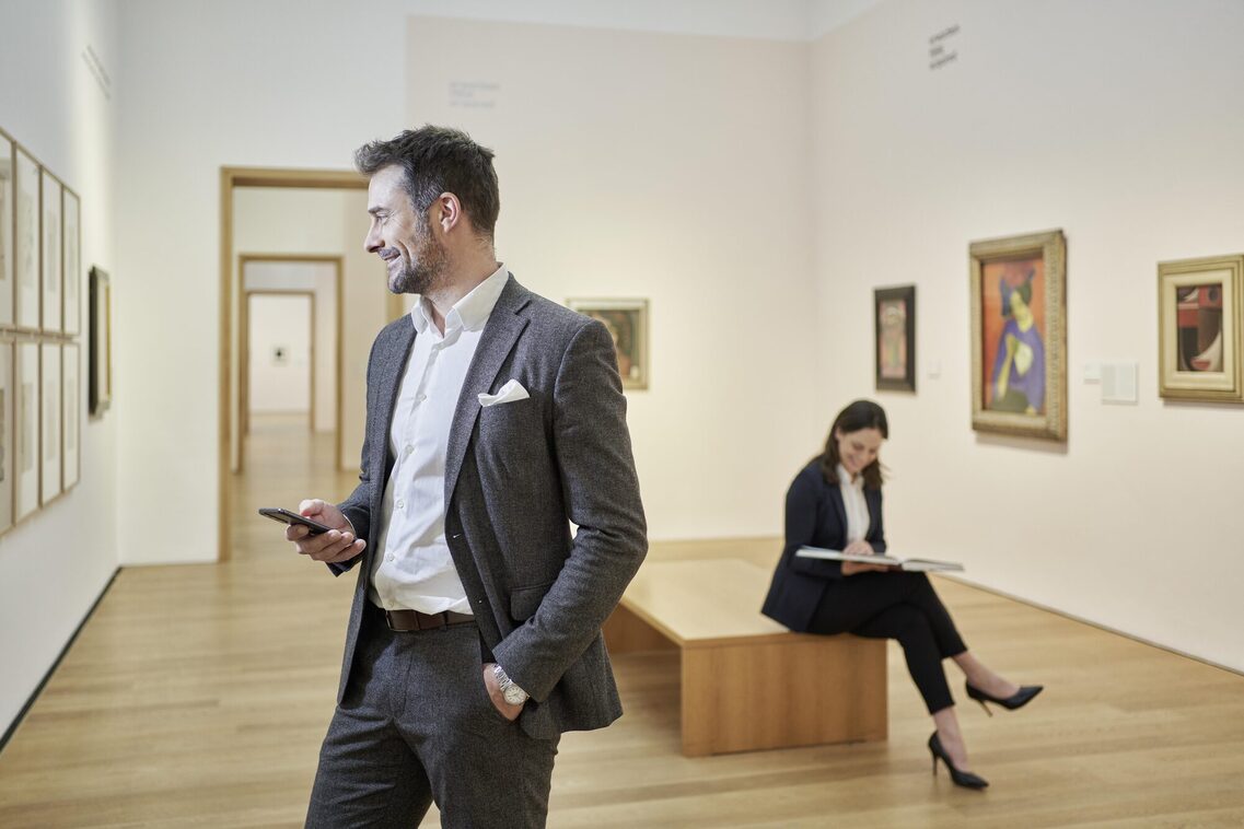 Ein Mann hält ein Handy und schaut sich ein Gemälde im Museum an, eine Frau im Hintergrund sitzt und liest ein Buch